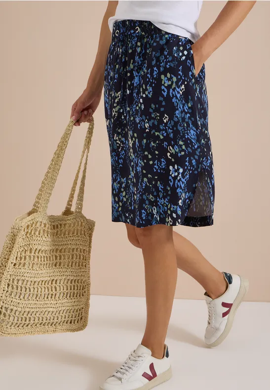 Vrouw in wit topje, blauw gebloemde midirok, witte sneakers en rieten tas voor een beige achtergrond.