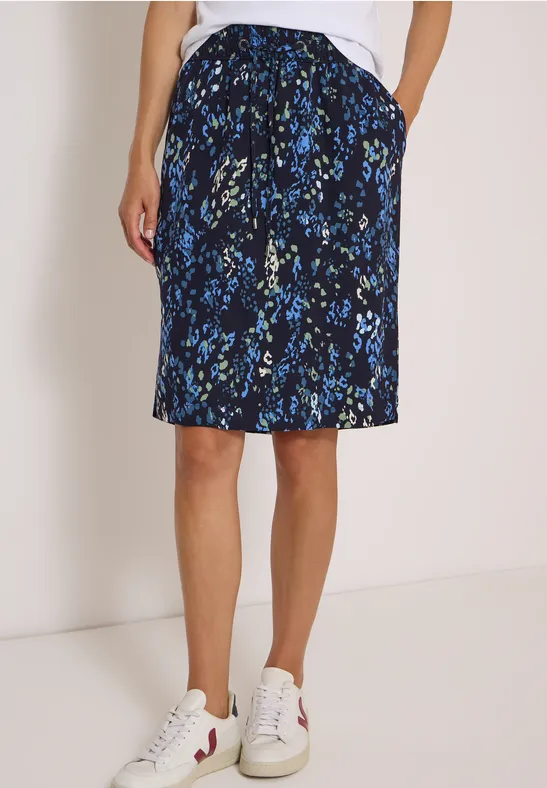 Vrouw draagt een zwarte knielange rok met blauw bloemenpatroon en zakken, gecombineerd met witte sneakers.