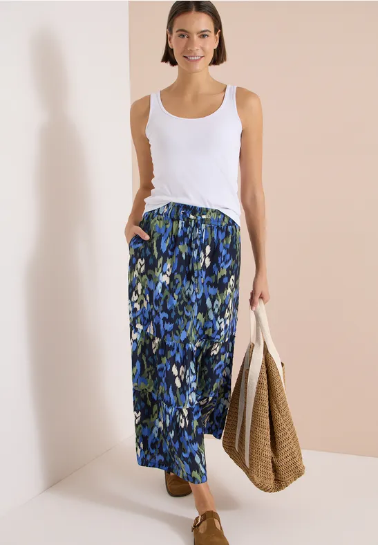Dame trägt weißes Top, blauen gemusterten Maxirock und hält eine geflochtene Tasche.