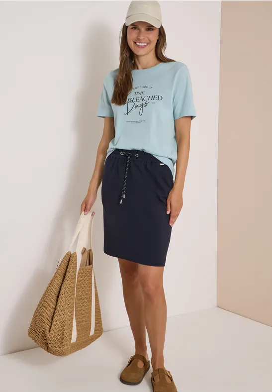 Frau trägt hellblaues T-Shirt, dunkelblauen Rock, Strohtasche, Cap und braune Schuhe, lächelnd.