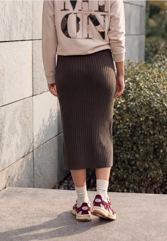 Person von hinten in beigefarbenem Pullover, dunkelbraunem Midirock, weißen Socken und roten Sneakern draußen.
