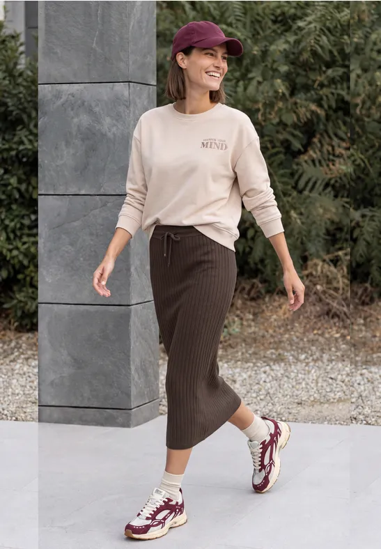 Eine lächelnde Frau spaziert draußen in einem lässigen Outfit mit Pullover, langer Rock, Cap und Sneakern.