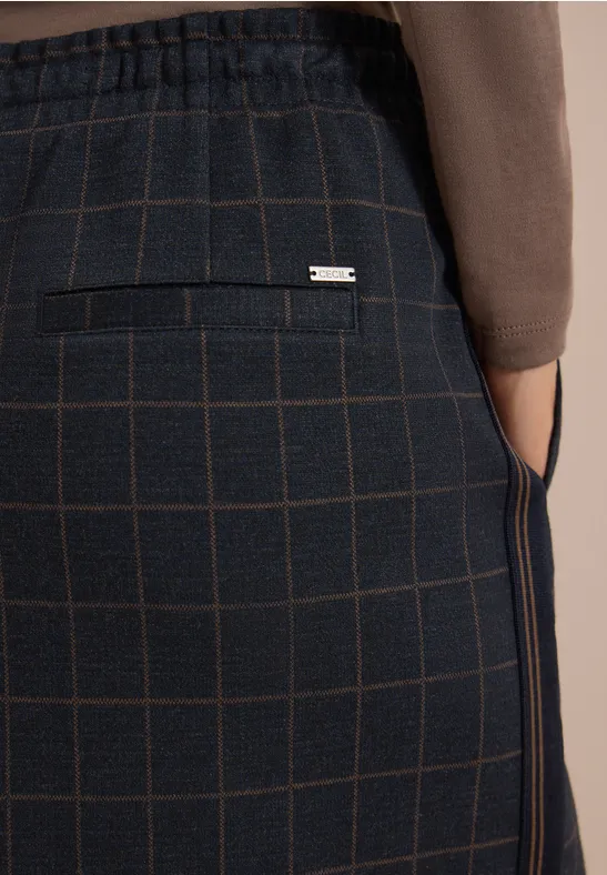 Detailaufnahme einer dunkelkarierten Hose mit Gesäßtasche und silbernem Label, Person trägt braunen Pullover.