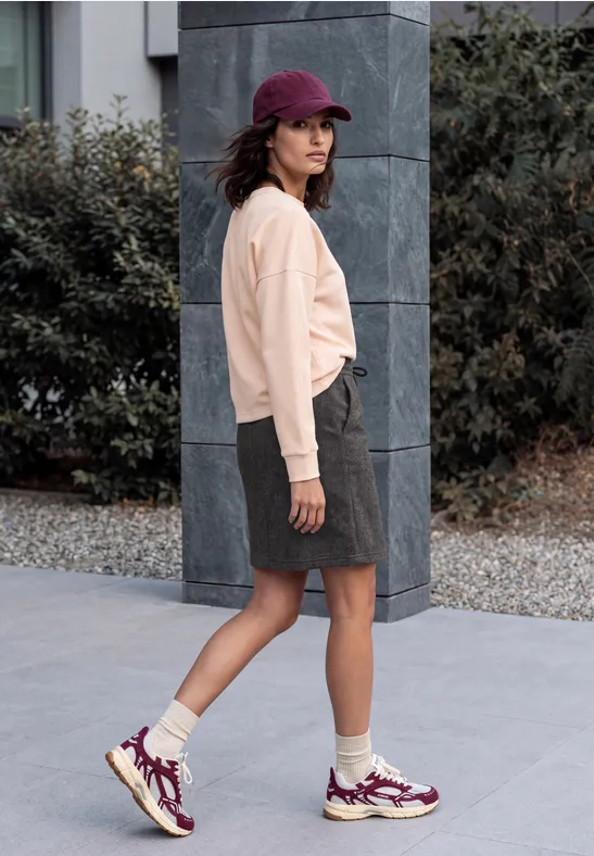 Vrouw in beige sweater, grijze rok, paarse sneakers en pet loopt buiten langs een zuil.