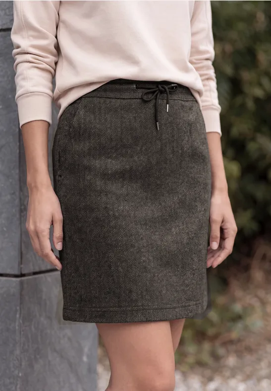 Iemand draagt een beige trui en een donkergroene wollen rok, staand naast een betonnen muur.