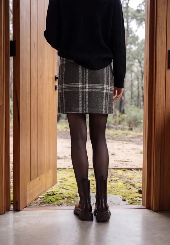 Frau steht in offener Holztür, trägt karierten Rock, schwarze Strumpfhose und Boots, Blick auf bewaldeten Hof.