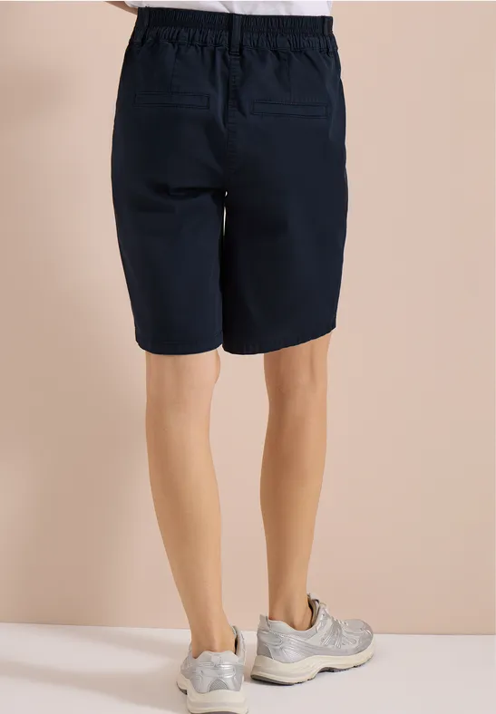 Achteraanzicht van persoon in donkerblauwe knielange short en lichte sneakers voor beige achtergrond.