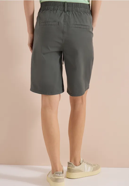 Achteraanzicht van persoon in grijze korte broek en lichte sneakers tegen een beige achtergrond.