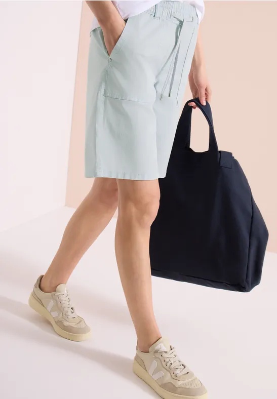 Een persoon draagt lichtgroene shorts, beige sneakers en houdt een grote zwarte tas vast.