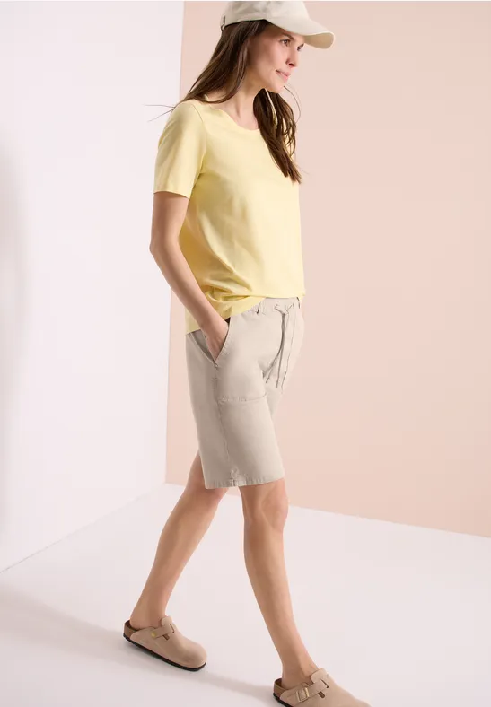 Vrouw in geel shirt, beige korte broek en instappers tegen lichte achtergrond.