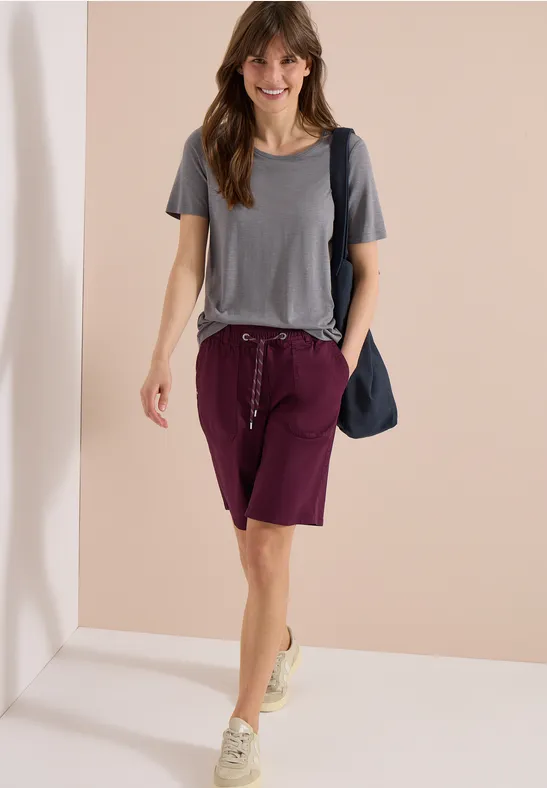 Een vrouw in grijs t-shirt, wijnrode bermuda's en sneakers, loopt met een blauwe schoudertas.