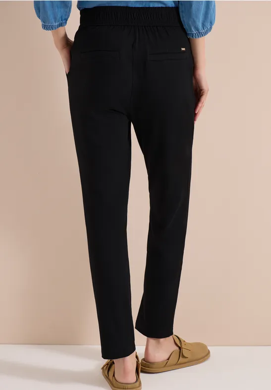 Person in schwarzen Hosen, blauem Oberteil und beigen Loafers vor schlichtem Beige-Hintergrund.