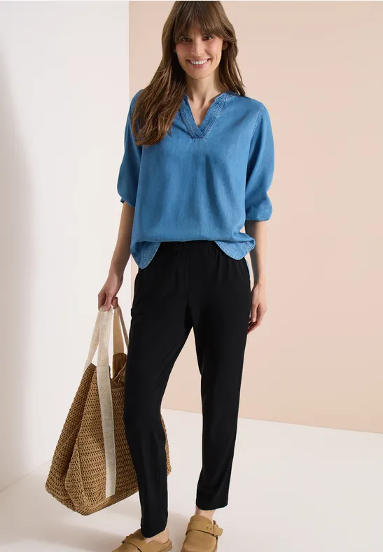 Frau trägt lockeres blaues V-Shirt, schwarze Hose und eine geflochtene Strohtasche vor neutralem Hintergrund.