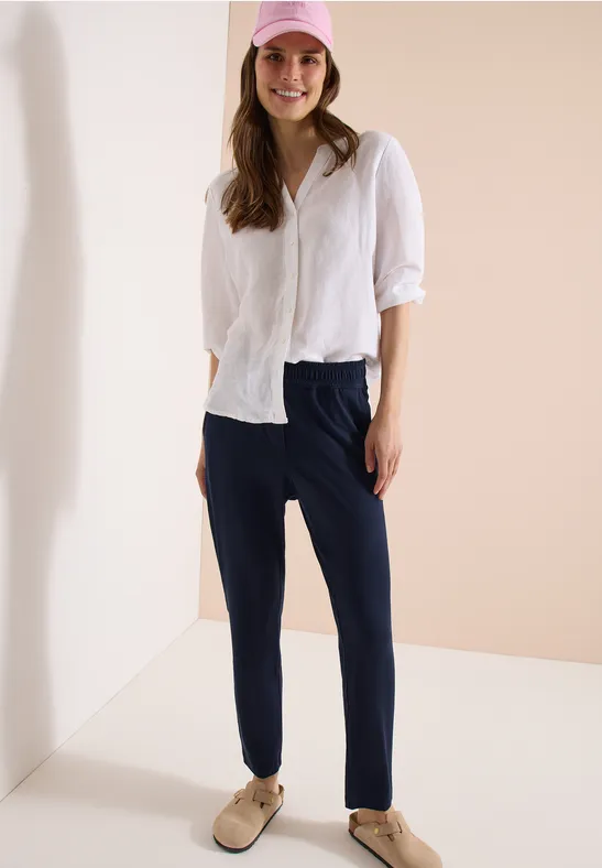 Frau mit weißem Hemd und dunklen Hosen steht vor hellem Hintergrund.