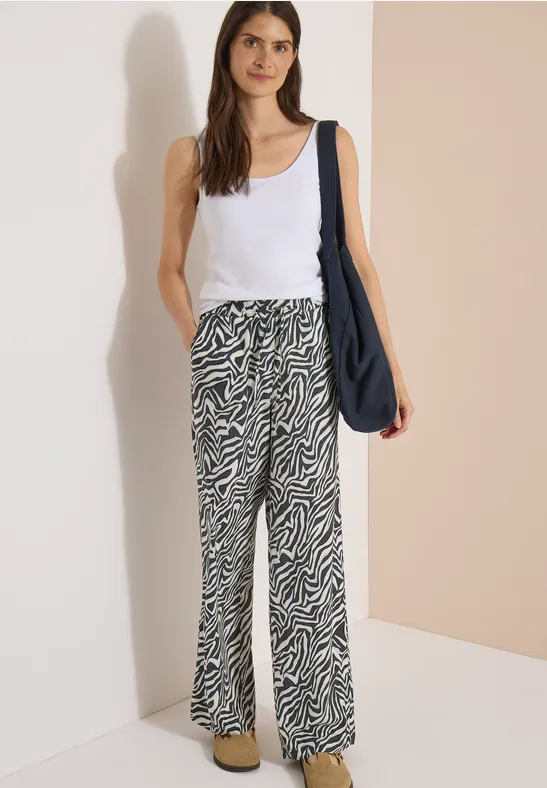 Femme en débardeur blanc, pantalon large à motif zébré et sac bandoulière bleu marine, debout devant un mur beige.