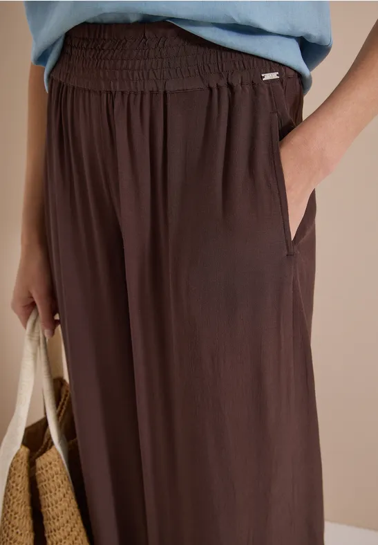 Nahaufnahme einer braunen, locker sitzenden Hose mit Gummibund und Seitentaschen, Hand in Tasche, Strohtasche am Arm.