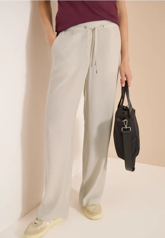 Personne porte un pantalon large en lin clair à cordon, des baskets crème et tient un sac noir.