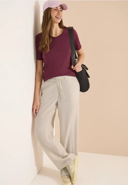 Femme décontractée: T-shirt bordeaux, pantalon large clair, baskets jaunes, sac noir et casquette rose.