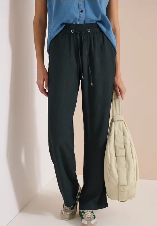 Schwarze, weite Hose mit Kordelzug, blaues Top, hält beige Steppjacke, kombiniert mit Sneakern.