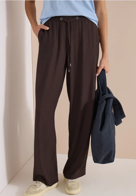 Frau trägt weite, dunkelbraune Hose, hellblaues Oberteil, weiße Sneaker, hält lässig eine dunkelblaue Tasche.