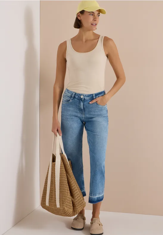 Vrouw met wit hemdje, opgerolde jeans, beige pet en sneakers, met bijpassende tas, voor een eenvoudige achtergrond.
