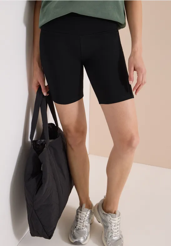 Frau trägt schwarze Radlerhose, grünes Oberteil, silberne Sneaker und hält eine große gesteppte schwarze Tasche.