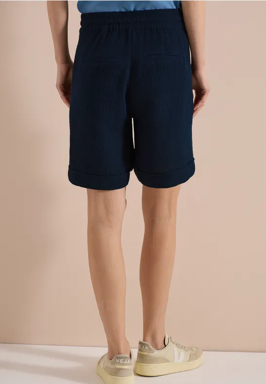 Ein Mann steht von hinten in dunkelblauen, knielangen Shorts und beigen Freizeitschuhen.