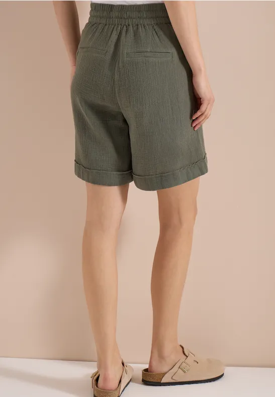 Rückansicht einer Frau in khaki Shorts und beigen Pantoletten vor schlichtem Hintergrund.