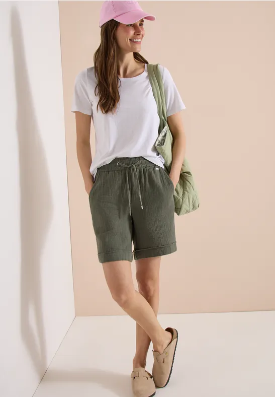 Eine junge Frau trägt ein weißes T-Shirt, khakigrüne Shorts, Sandalen, eine rosa Kappe und eine grüne Tasche.