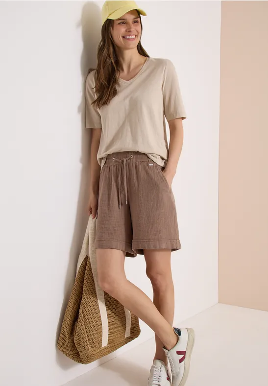 Frau lehnt an Wand mit beiger Bluse, braunen Shorts, weißen Sneakern und geflochtener Tasche.