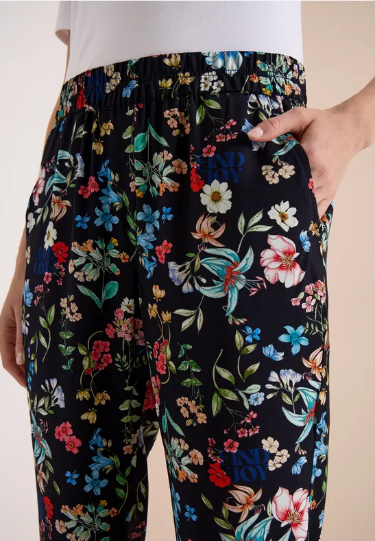 Schwarze, locker sitzende Hose mit buntem Blumenmuster, Hand in seitlicher Tasche.