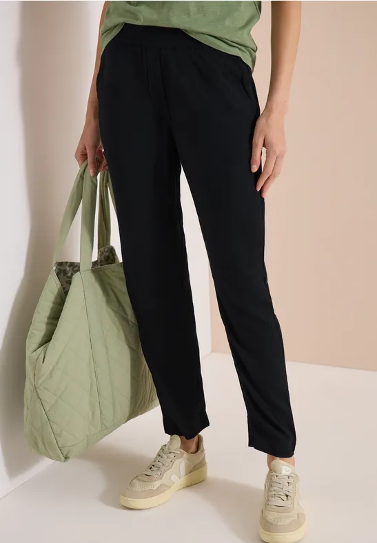 Persoon draagt donkerblauwe broek, groen shirt, heeft een doorgestikte groene shopper en beige sneakers.
