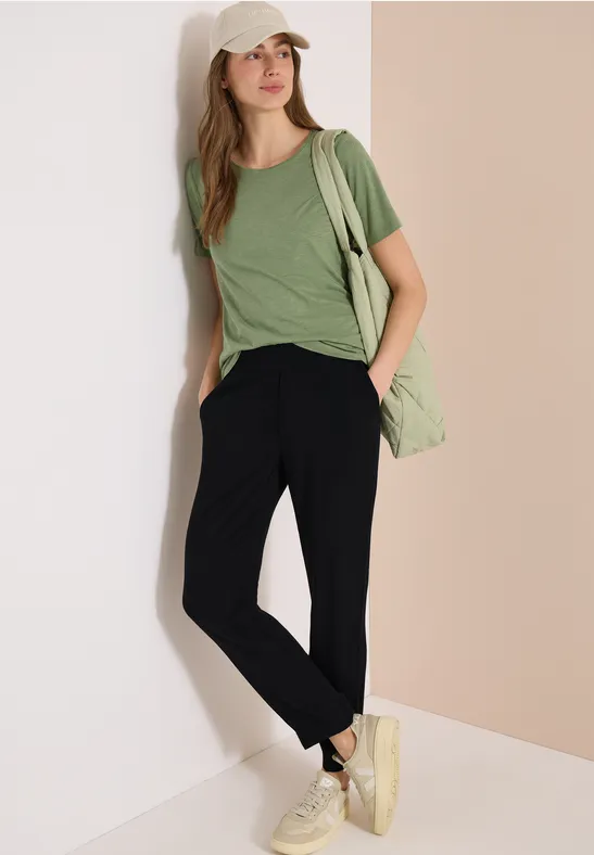 Een vrouw leunt tegen een muur, met een groen t-shirt, zwarte broek, witte schoenen en een groene tas.