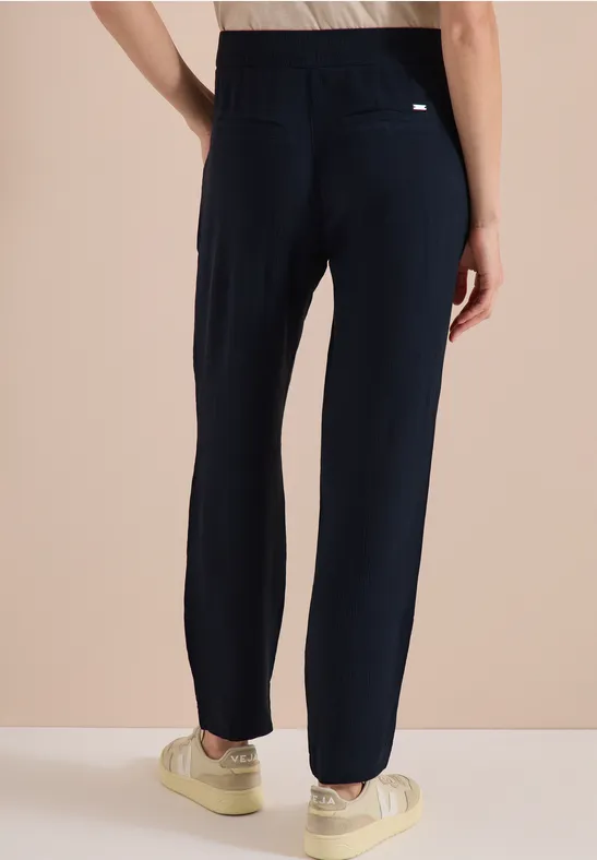 Persoon van achteren in donkerblauwe, rechte broek en crèmekleurige sneakers voor een beige achtergrond.