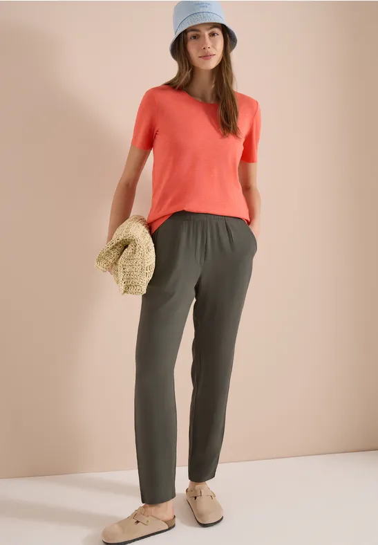 Vrouw in rood T-shirt, groene broek en beige schoenen houdt een strohoed vast voor een effen achtergrond.