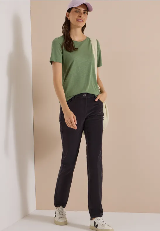 Femme en t-shirt vert, jean foncé, casquette beige et baskets blanches devant un mur beige.