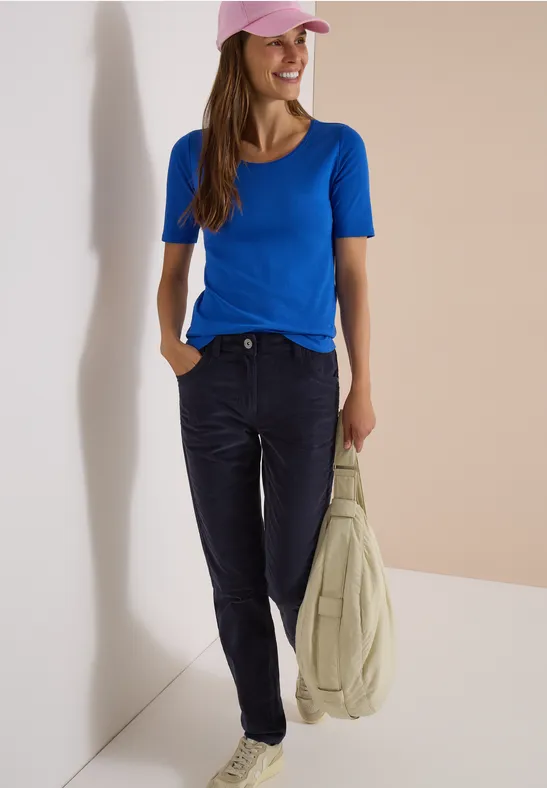 Eine Frau trägt ein blaues T-Shirt und eine rosa Kappe, hält eine helle Jacke vor neutralem Hintergrund.
