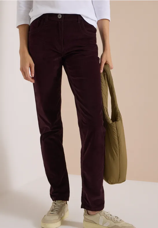Personne en pantalon en velours côtelé bordeaux, baskets blanches, sac kaki, fond beige clair.