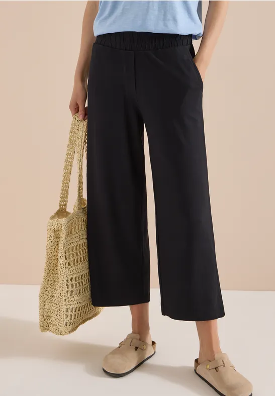 Schwarze, weit geschnittene Culotte mit hohem Bund, kombiniert mit Basttasche und beigen Slippern.