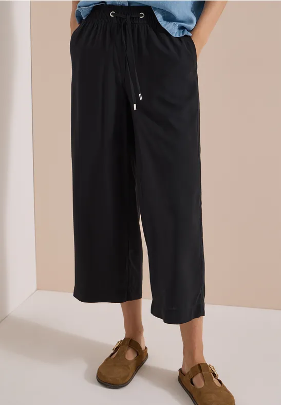Schwarze, weite Culotte-Hose mit Kordelzug und braunen Sandalen vor neutralem Hintergrund