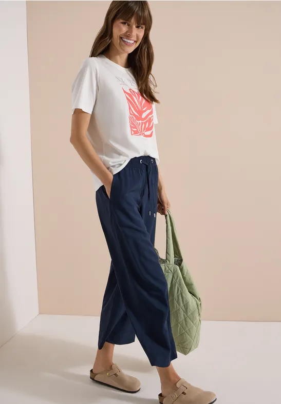 Frau in weißem T-Shirt mit rotem Print, blauen weiten Hose, Sandalen und grünem Shopper.
