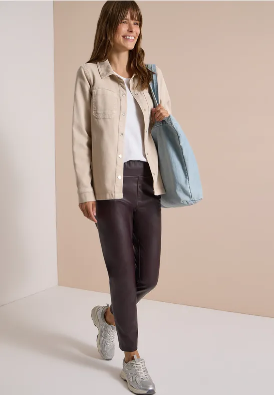 Frau in beiger Jacke, weißem Shirt, dunklen Leggings, Sneakern und großer hellblauer Tasche, die lächelnd posiert.