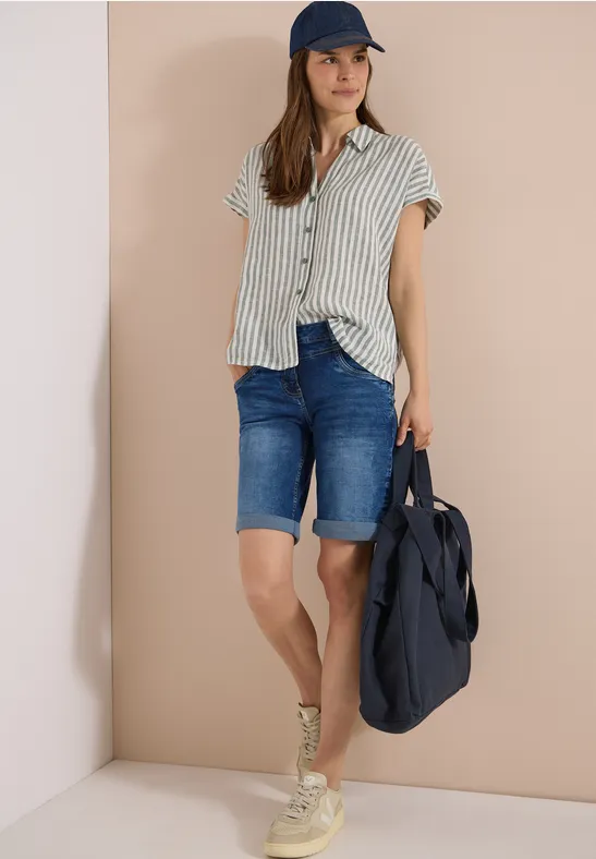 Frau mit gestreiftem Kurzarmhemd, Jeans-Bermudas, Sneakers, Baseballcap und großer blauer Tasche.