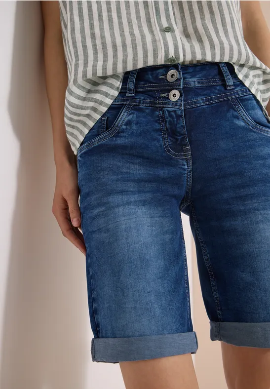 Frau in gestreiftem Top und hochgekrempelter Jeans-Shorts vor weißer Wand