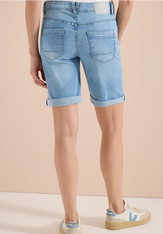 Person von hinten in hellblauen Jeans-Shorts mit Umschlag und beigen Sneakern vor hellem Hintergrund