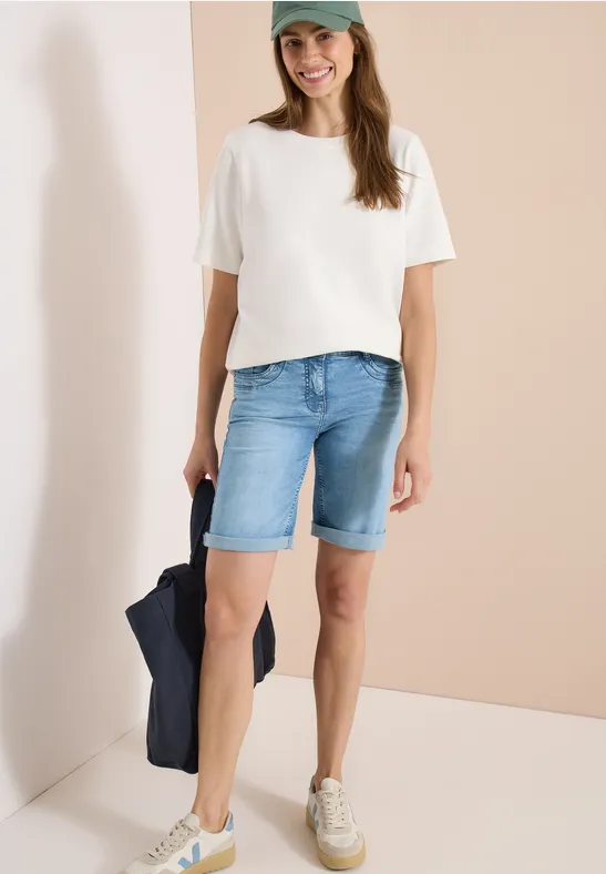 Frau trägt weiße T-Shirt, blaue Jeans-Bermudas und weiße Sneaker.