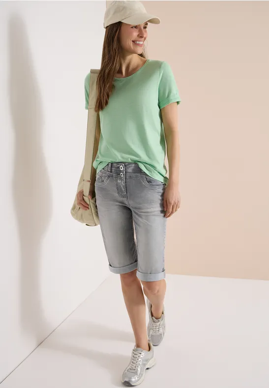 Een vrouw in een groen t-shirt, knielange spijkershort, sneakers, met strooien tas en pet voor neutrale achtergrond.