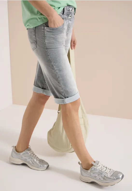 Benen in lichtblauwe, omgeslagen bermudashorts, zilveren sneakers, groen topje en beige canvas tas.