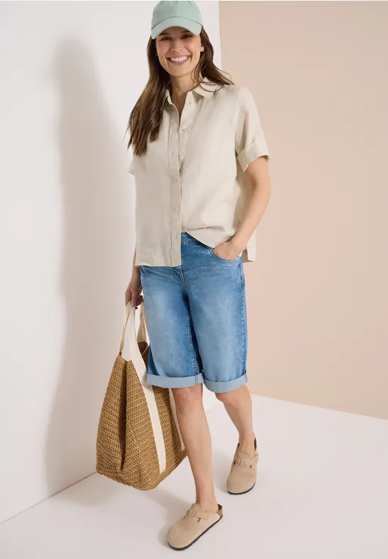 Frau im beigen Kurzarmhemd und Jeans-Bermuda, mit Strohtasche und Espadrilles vor zweifarbigem Hintergrund.