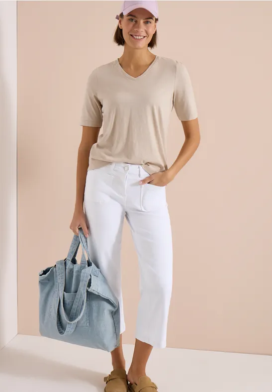 Vrouw in beige t-shirt, witte capri-jeans en grote lichtblauwe shopper voor een neutrale achtergrond.